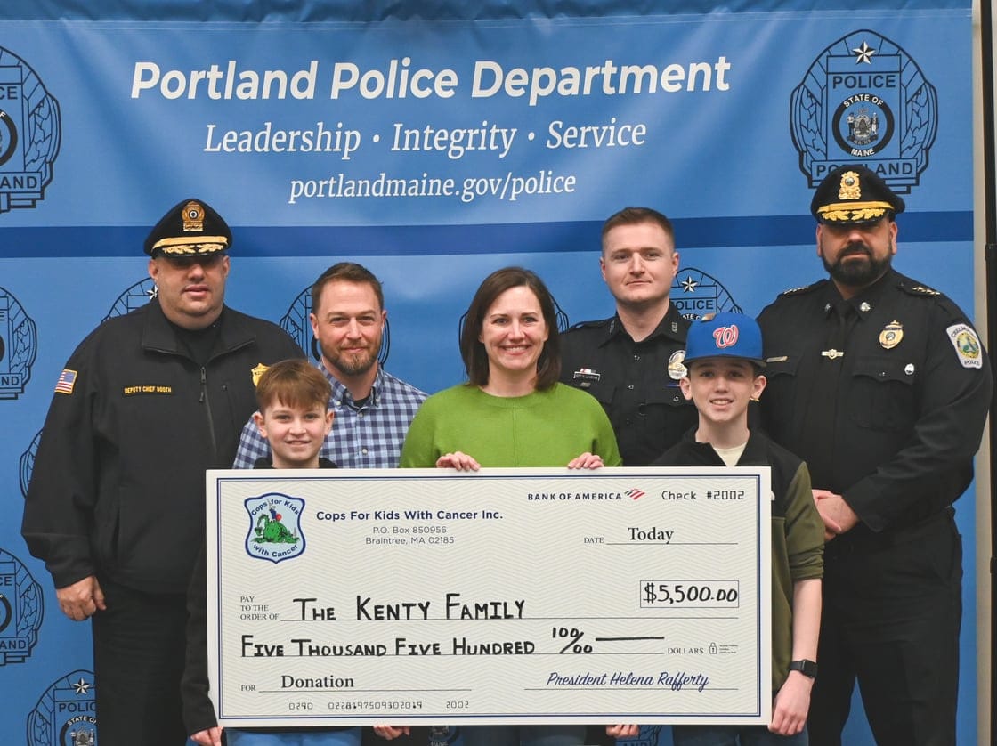 Cops for Kids with Cancer board members stand with the Kenty family of Portland, Maine, during a check presentation on Monday at the Portland (ME) Police Department. From left are Deputy Chief Andrew Booth of the Carlisle (MA) Police Department, James Kenty, Tim Kenty, Allison Kenty, Ryan Linehan of the Nashua (NH) Police Department, Will Kenty, and Deputy Chief Gary Hannagan of the Chelmsford (MA) Police Department.